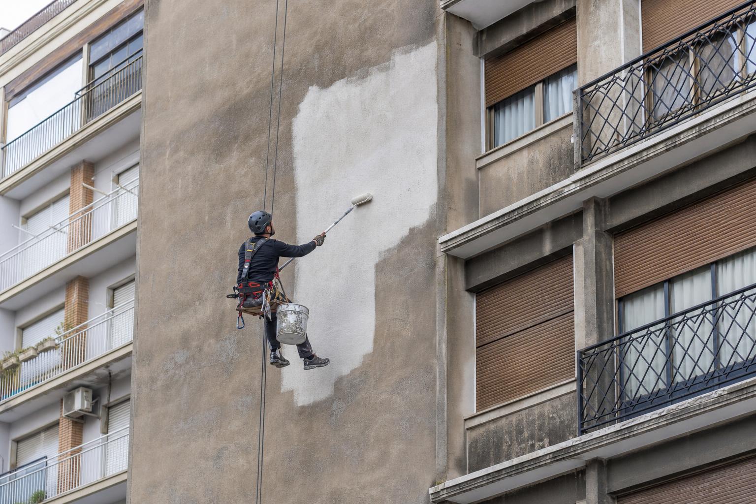 Operario rehabilitando y pintando la fachada de un edificio suspendido de cuerdas, sin andamios